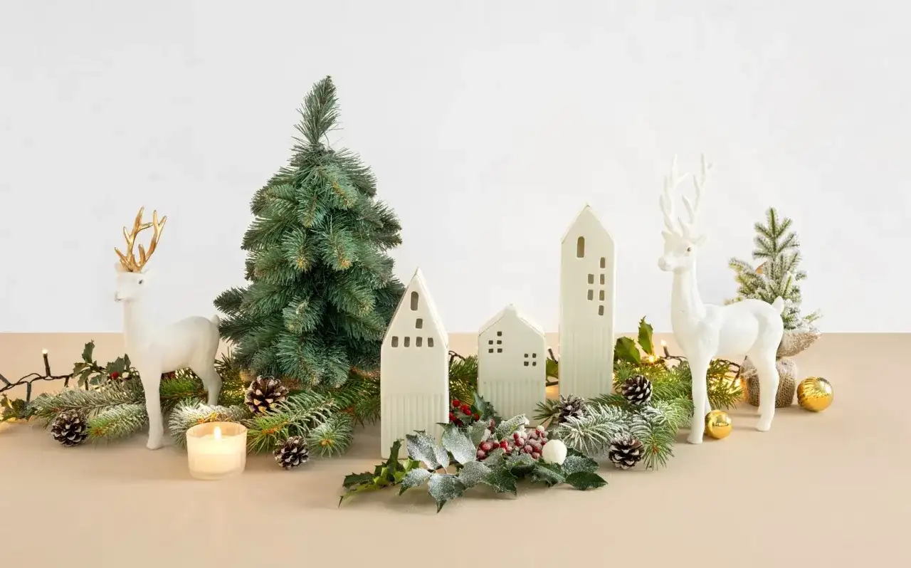 Bożonarodzeniowa dekoracja: białe renifery, choinka, ceramiczne domki, lampki i szyszki.