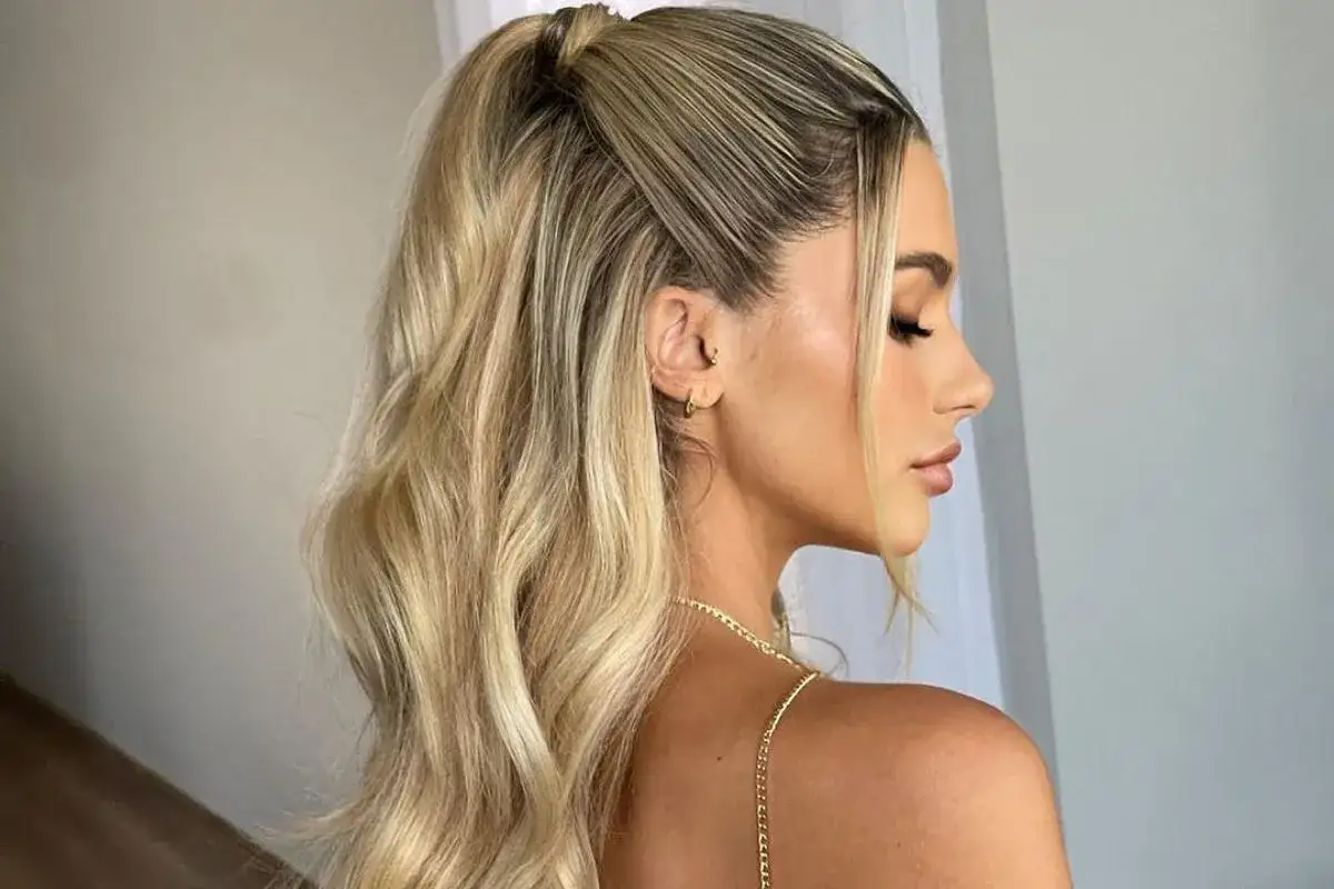 Eleganckie fryzury damskie na wesele: blond włosy upięte w wysoki kucyk z delikatnymi falami i dwoma pasmami opadającymi na twarz.