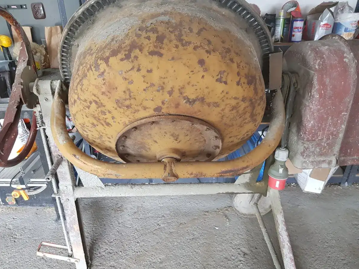 Stara betoniarka 150l, zardzewiała, wymaga wymiany łożysk. W tle widać narzędzia i materiały budowlane.