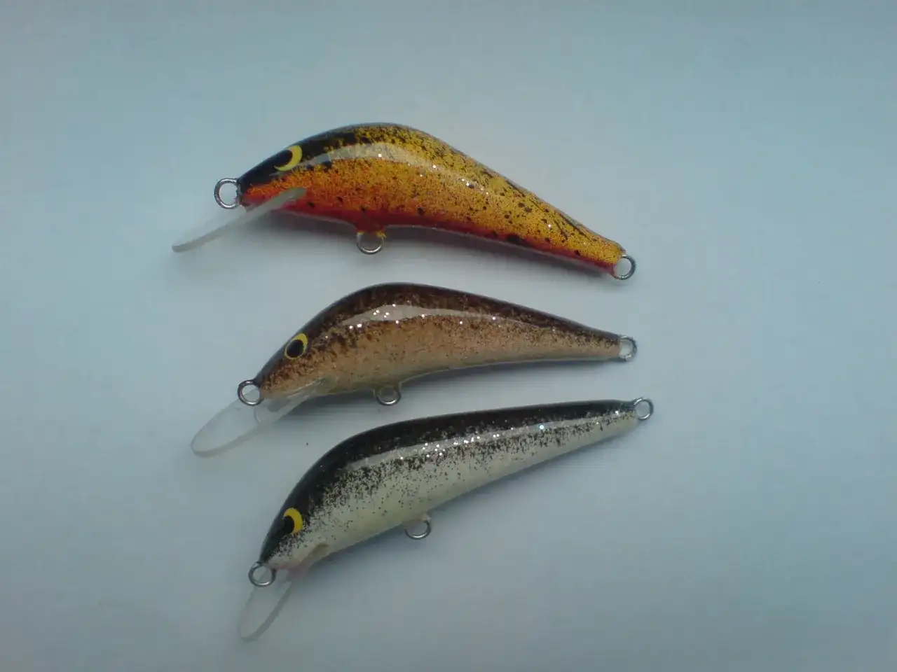 Trzy spinningowe przynęty na pstrąga w różnych kolorach: żółto-pomarańczowa, brązowa i czarno-biała.