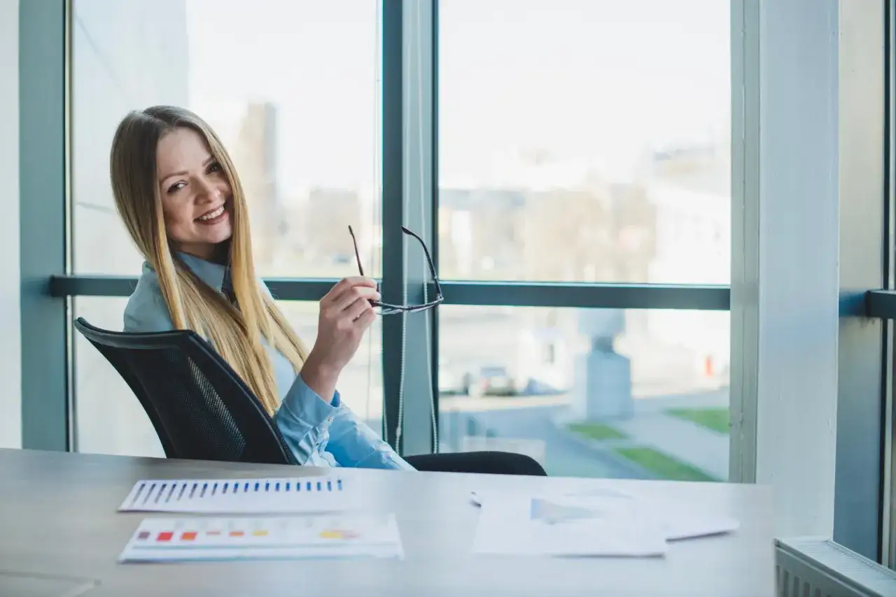 Uśmiechnięta blondynka w biurze, trzymająca okulary, zastanawia się, kiedy można wziąć urlop bezpłatny.