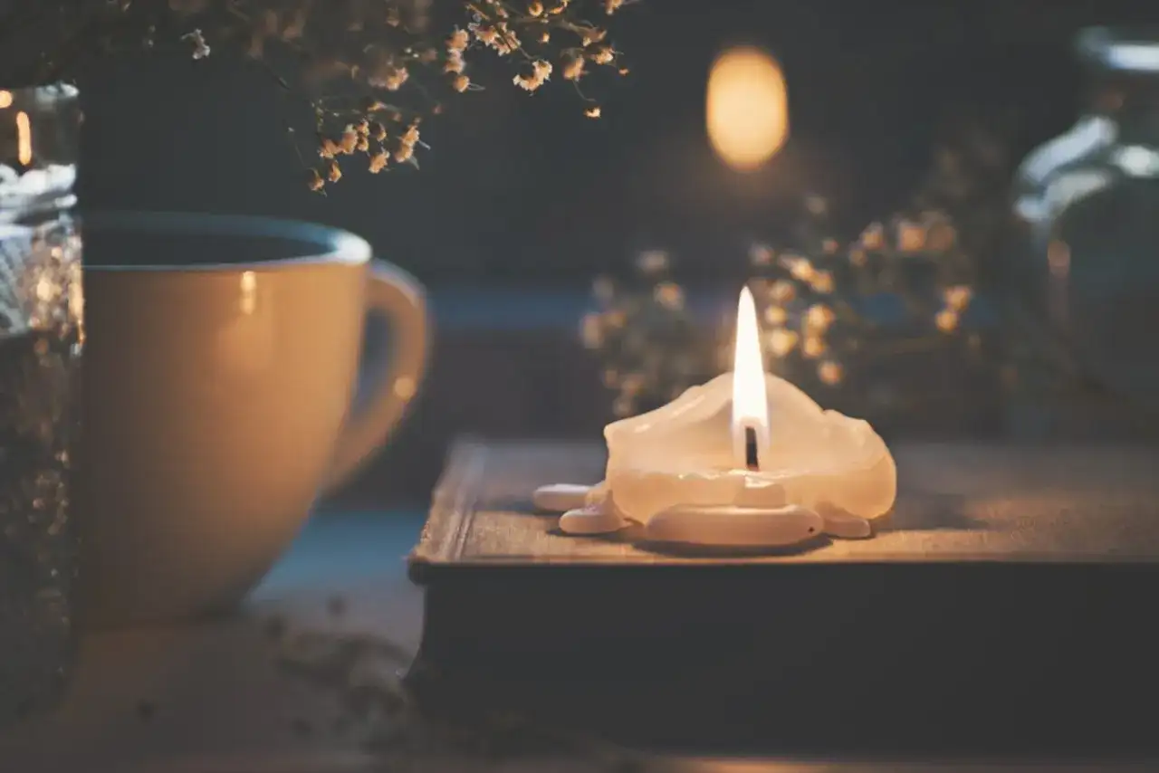 Płonąca świeca na książce, obok filiżanka i suszone kwiaty.
