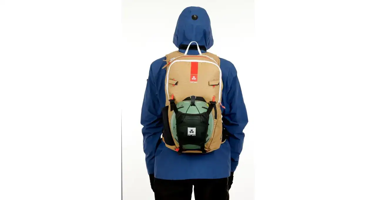 Osoba w niebieskiej kurtce ma na plecaku mocowanie kasku. Plecak jest beżowy z czerwonymi akcentami.