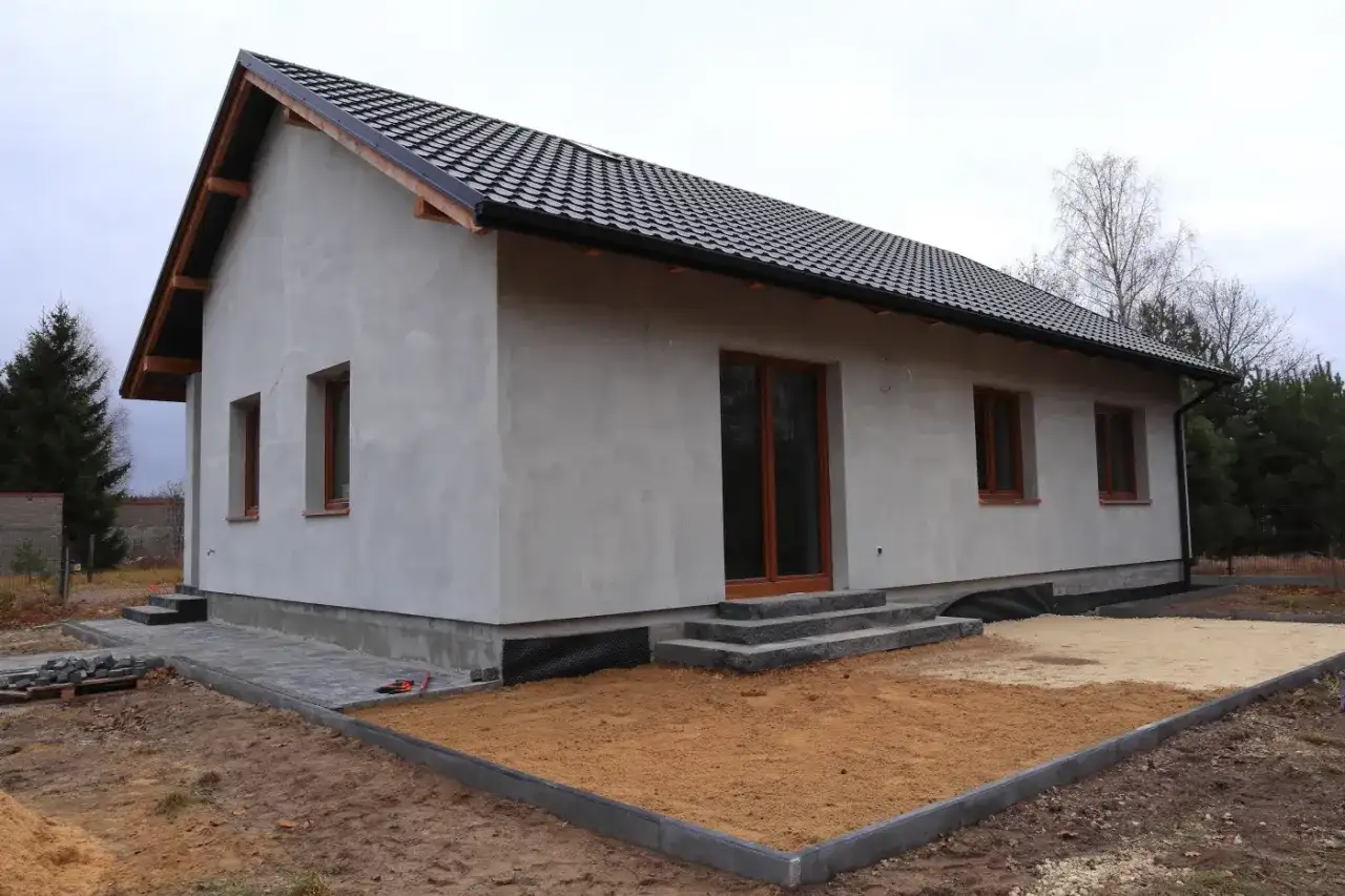 Nowy dom w trakcie budowy, z szarym tynkiem i brązowymi oknami. Wniosek o warunki zabudowy złożony, teren wokół domu przygotowany do dalszych prac.