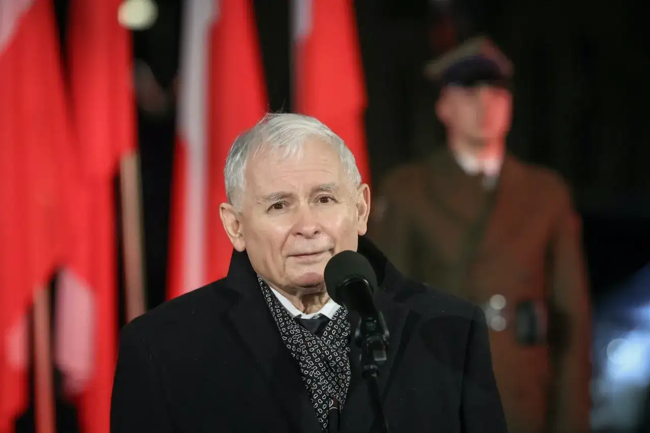 Jarosław Kaczyński przemawia przy mikrofonie na tle polskich flag i żołnierza.