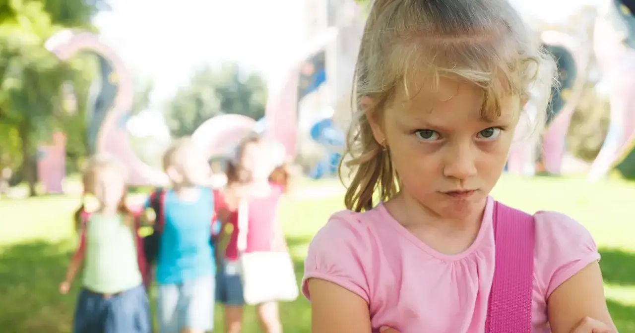 Zagniewana dziewczynka, bunt 7 latka, na tle bawiących się dzieci i placu zabaw.
