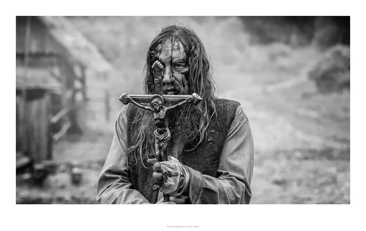 Mroczna recenzja książki "Malowany ptak". Postać z krucyfiksem, w brudnych ubraniach, z zakrwawioną twarzą.