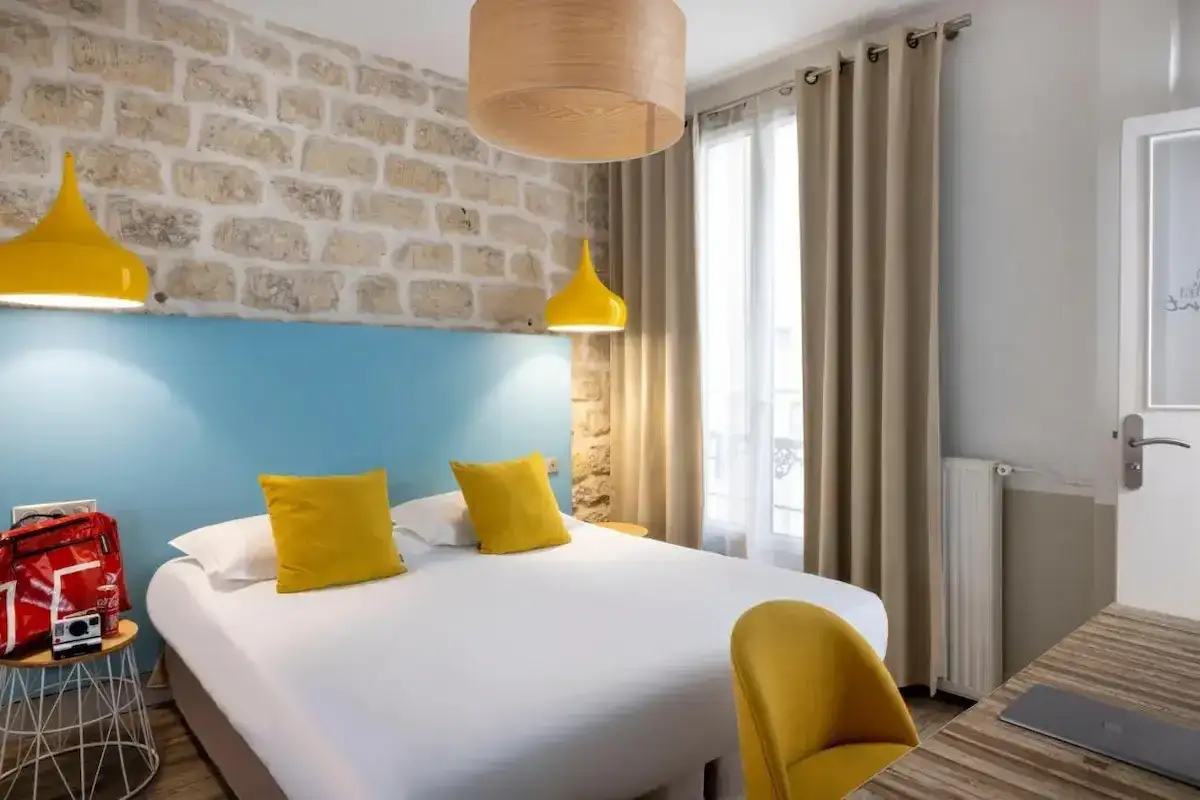 Przytulny pokój hotelowy z łóżkiem, żółtymi poduszkami i lampami, kamienną ścianą i oknem.