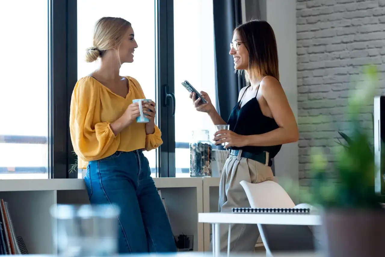 Dwie kobiety rozmawiają przy oknie, jedna z kubkiem, druga z telefonem. To przykład, jak wygląda **small talk co to** jest.