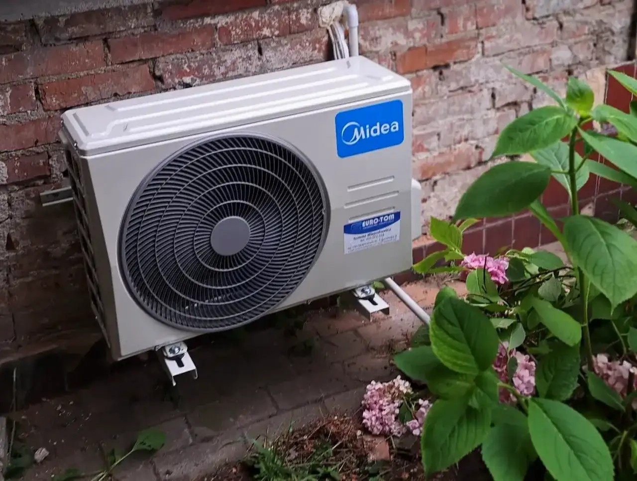 Jednostka zewnętrzna klimatyzatora Midea zamontowana na ceglanej ścianie, obok krzewu z różowymi kwiatami.