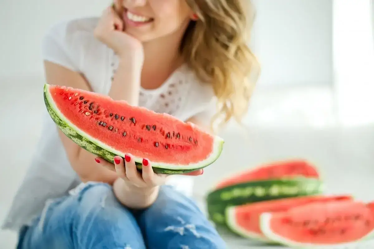 Uśmiechnięta kobieta trzyma kawałek arbuza, zastanawiając się, czy arbuz tuczy. W tle widać kolejne plastry soczystego owocu.