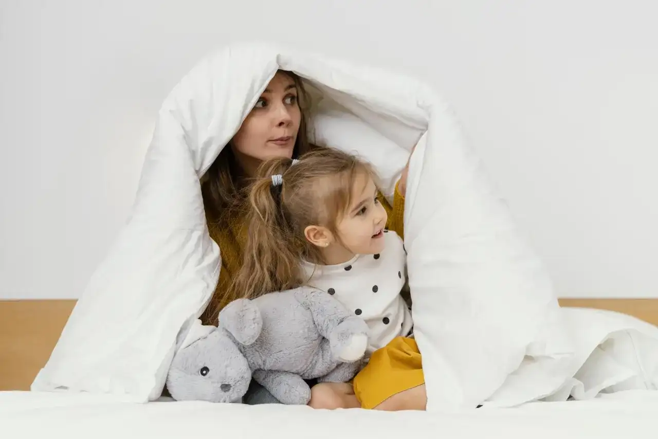 Mama i dziecko bawią się pod białą kołdrą, która przypomina przytulne kołderki dla niemowląt. Obok leży szary pluszowy króliczek.