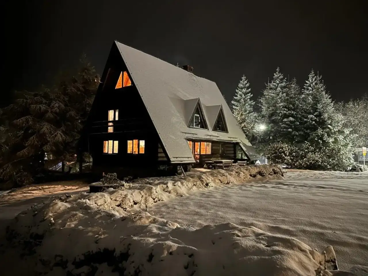 Drewniany domek oświetlony ciepłym światłem, otoczony śniegiem i choinkami. Idealne miejsce na zimowy wypoczynek na przełęczy okraj.