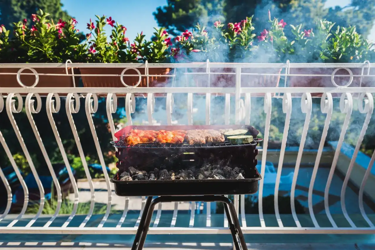 Grill z mięsem i warzywami na balkonie, w tle kwiaty i błękitne niebo.