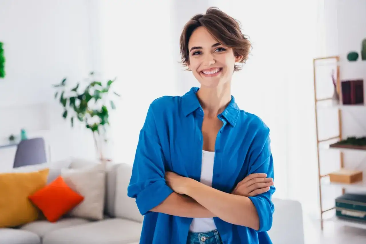 Uśmiechnięta kobieta w niebieskiej koszuli stoi ze skrzyżowanymi rękami w jasnym wnętrzu.