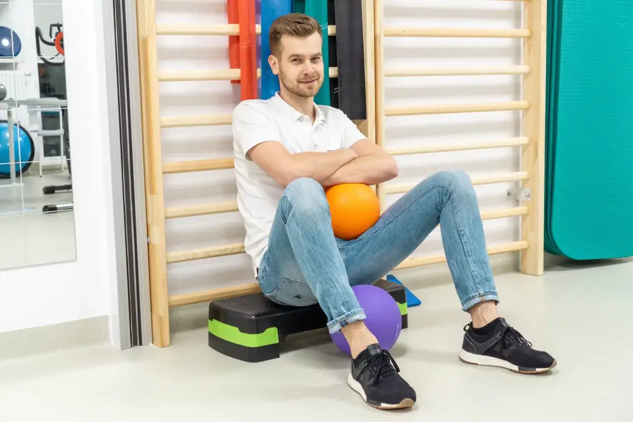 Fizjoterapeuta siedzi na schodku z piłkami, na tle drabinek i sprzętu rehabilitacyjnego.