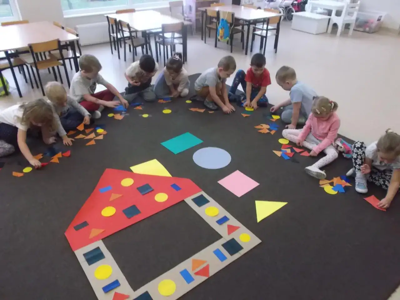 Dzieci w przedszkolu tworzą obrazki z kolorowych figur geometrycznych.