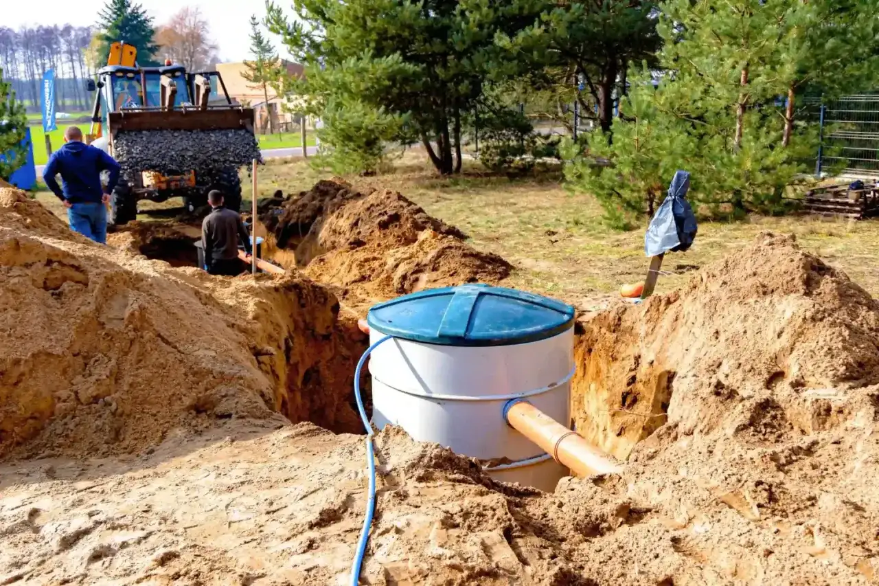 Trwa montaż oczyszczalni przydomowej. Koparka zasypuje wykop, a pracownicy nadzorują instalację zbiornika.