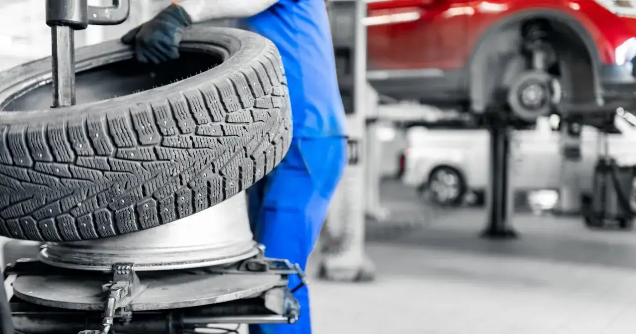 Mechanik w niebieskim kombinezonie trzyma oponę na tle warsztatu samochodowego.