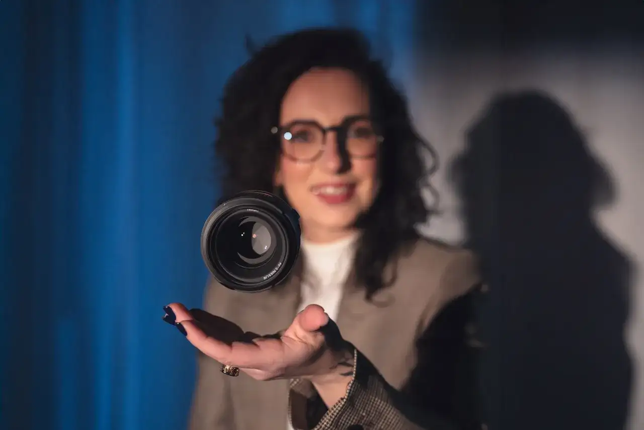 Uśmiechnięta kobieta w okularach unosi obiektyw aparatu nad dłonią. Czy fotograf przechowuje zdjęcia w obiektywie?