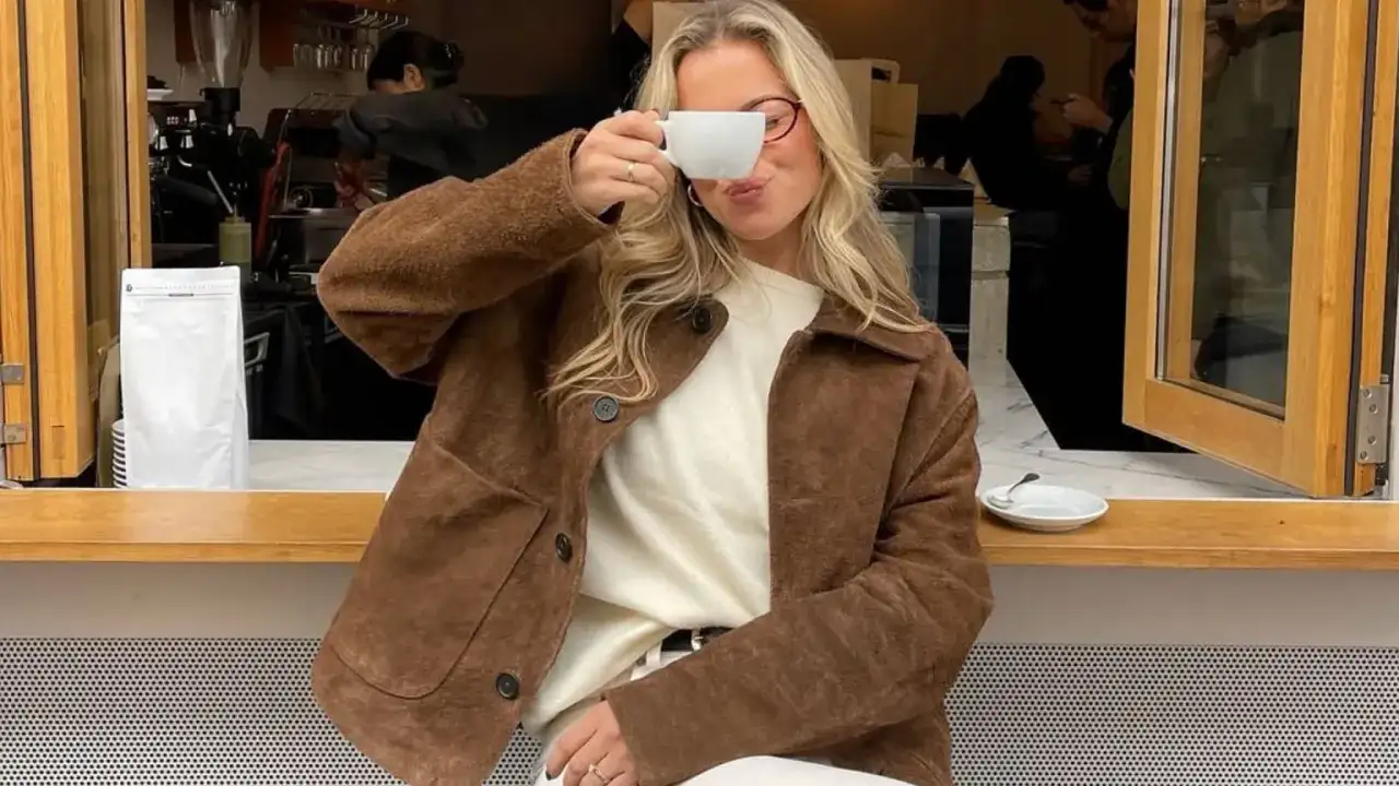Kobieta w brązowej kurtce zamszowej popija kawę w kawiarni.