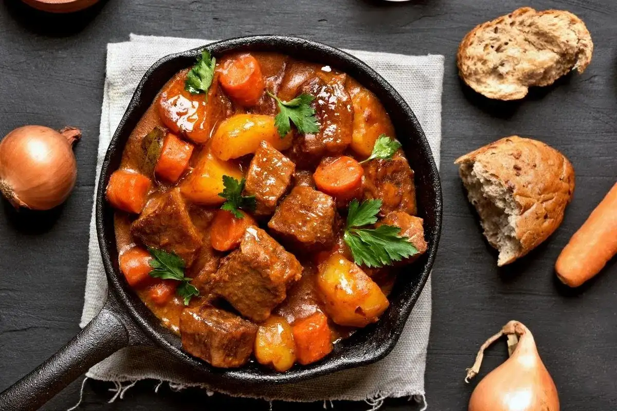 Gulasz wołowy z marchewką i ziemniakami w żeliwnej patelni. Ile kostek rosołowych na litr wody? Obok cebula, marchewka i chleb.