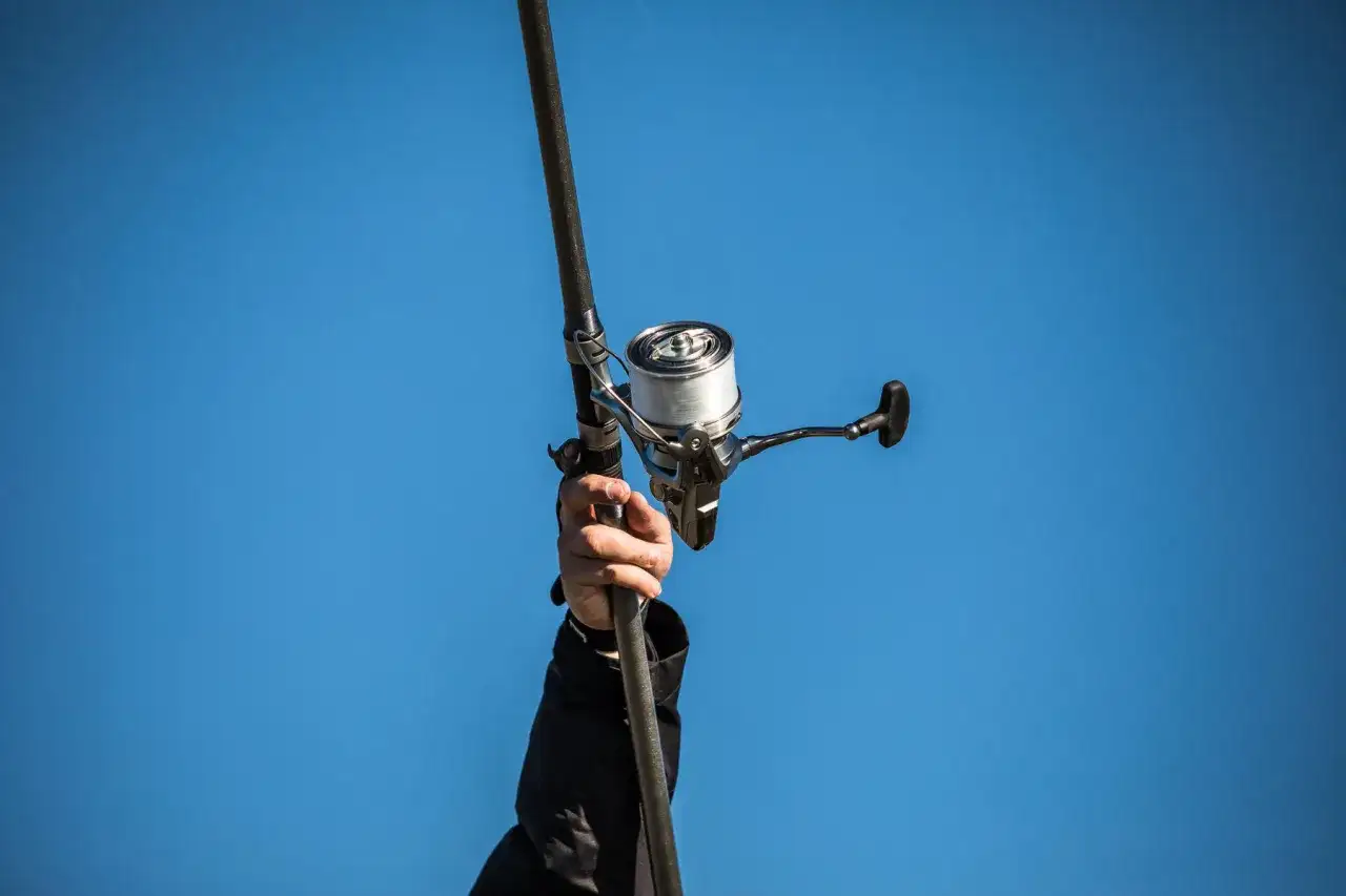 Dłoń trzyma wędkę z nawiniętą plecionką na kołowrotek, na tle błękitnego nieba.