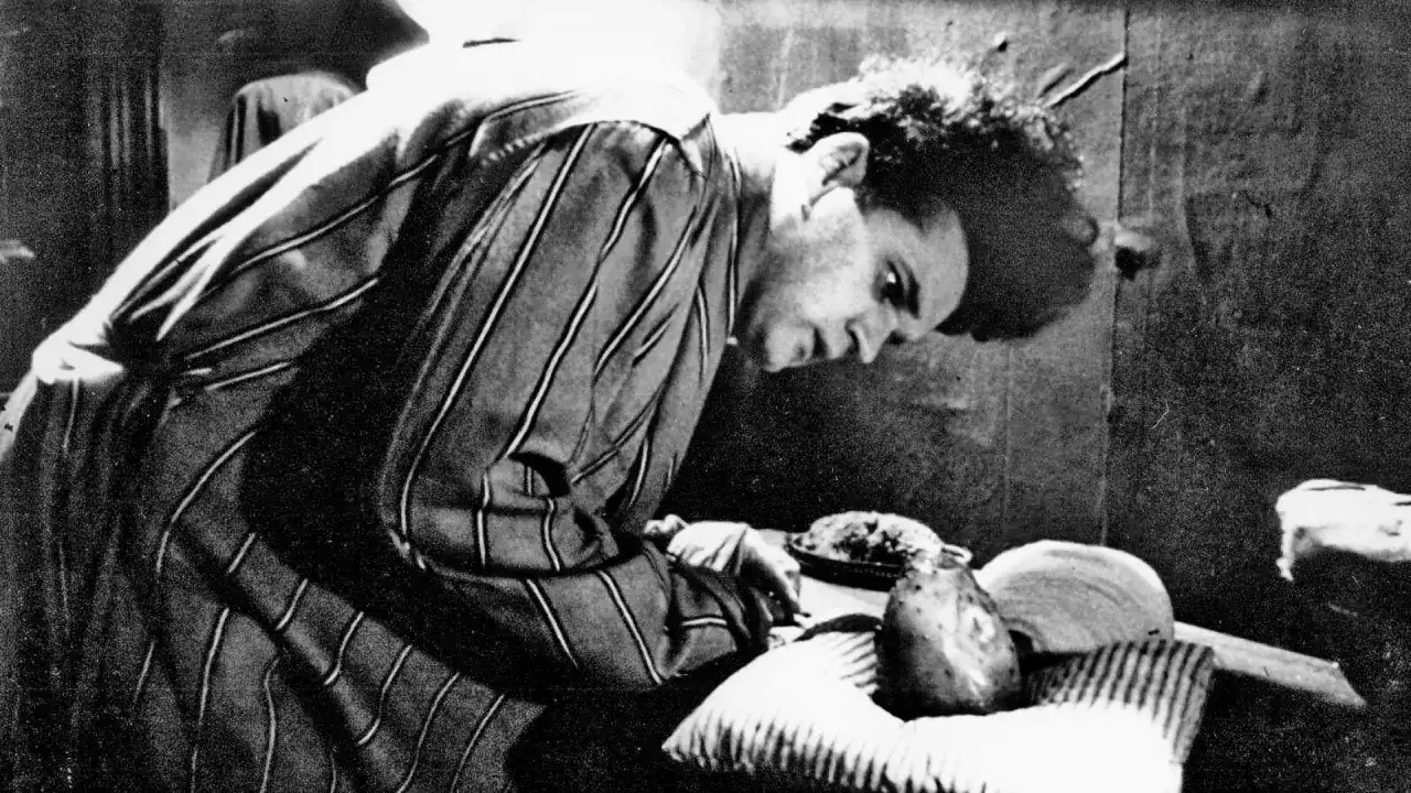 Młody David Lynch w pasiastym szlafroku pochyla się nad stołem, na którym leżą dziwne przedmioty.