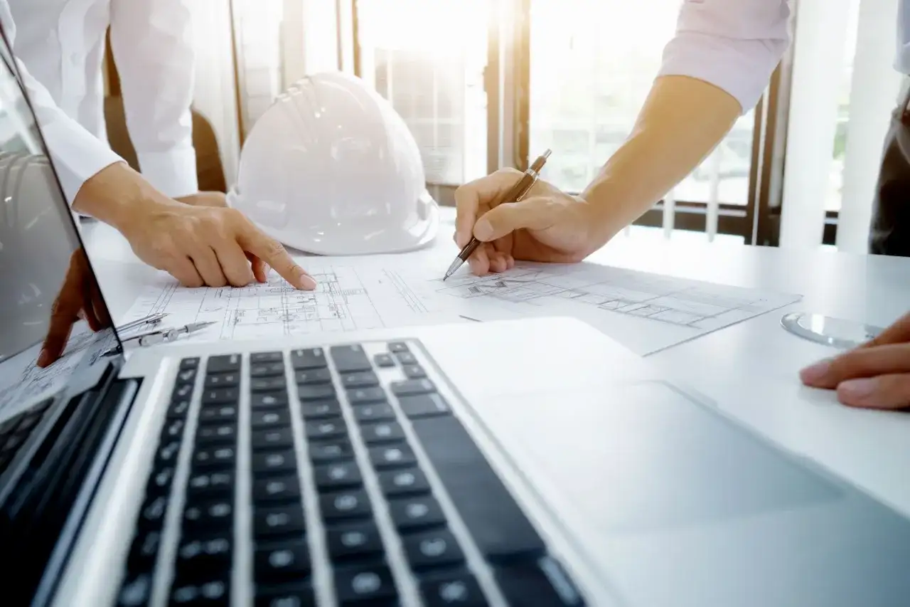 Inżynierowie analizują plany budowlane podczas rocznego przeglądu budowlanego. Na stole leży biały kask, laptop i dokumenty.