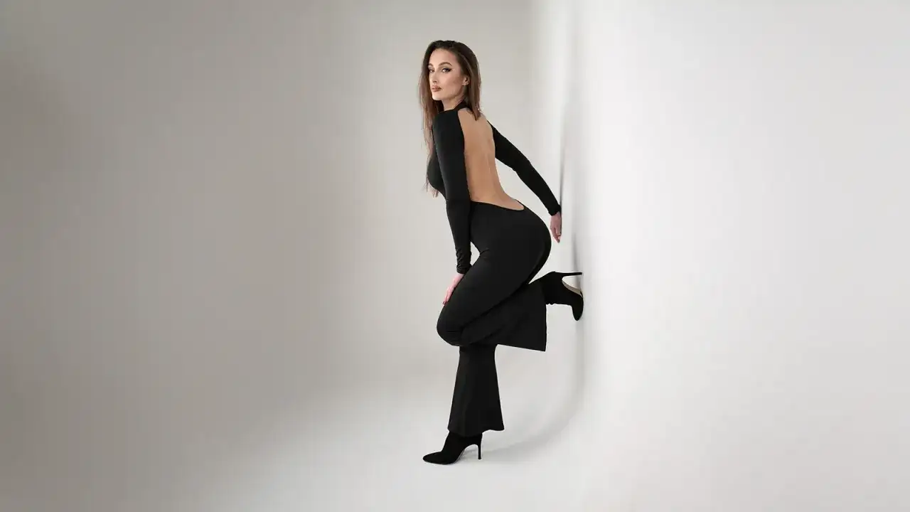 Modelka w czarnym, wyciętym z tyłu kombinezonie i butach na obcasie opiera się o białą ścianę.