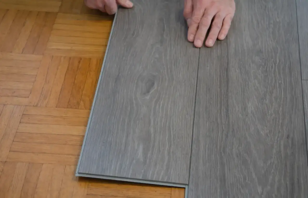 Ręce układają szare panele winylowe na starym parkiecie. Czy można kłaść panele na panele? To pytanie, na które odpowiada ta wizualizacja.