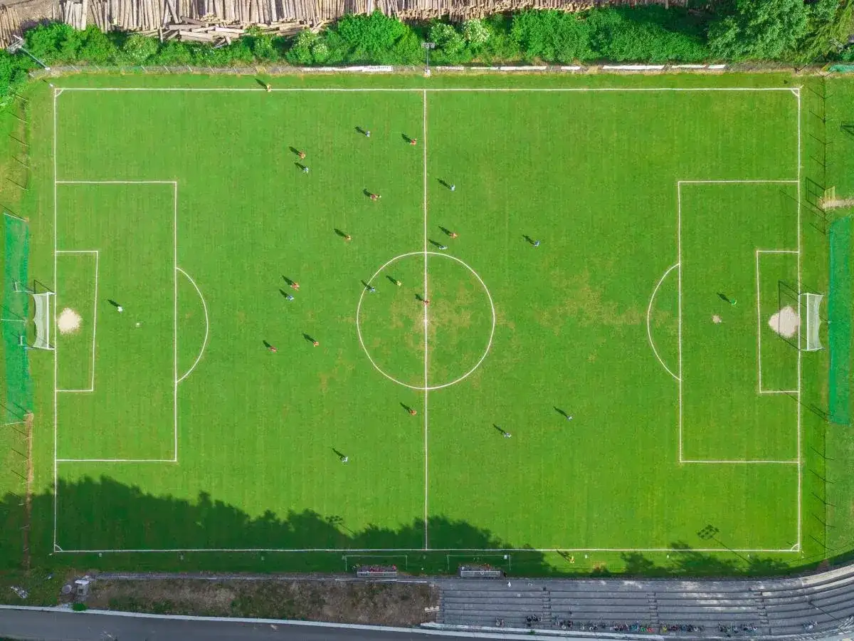 Widok z lotu ptaka na zielone boisko piłkarskie z zaznaczonymi liniami, bramkami i kilkoma zawodnikami.