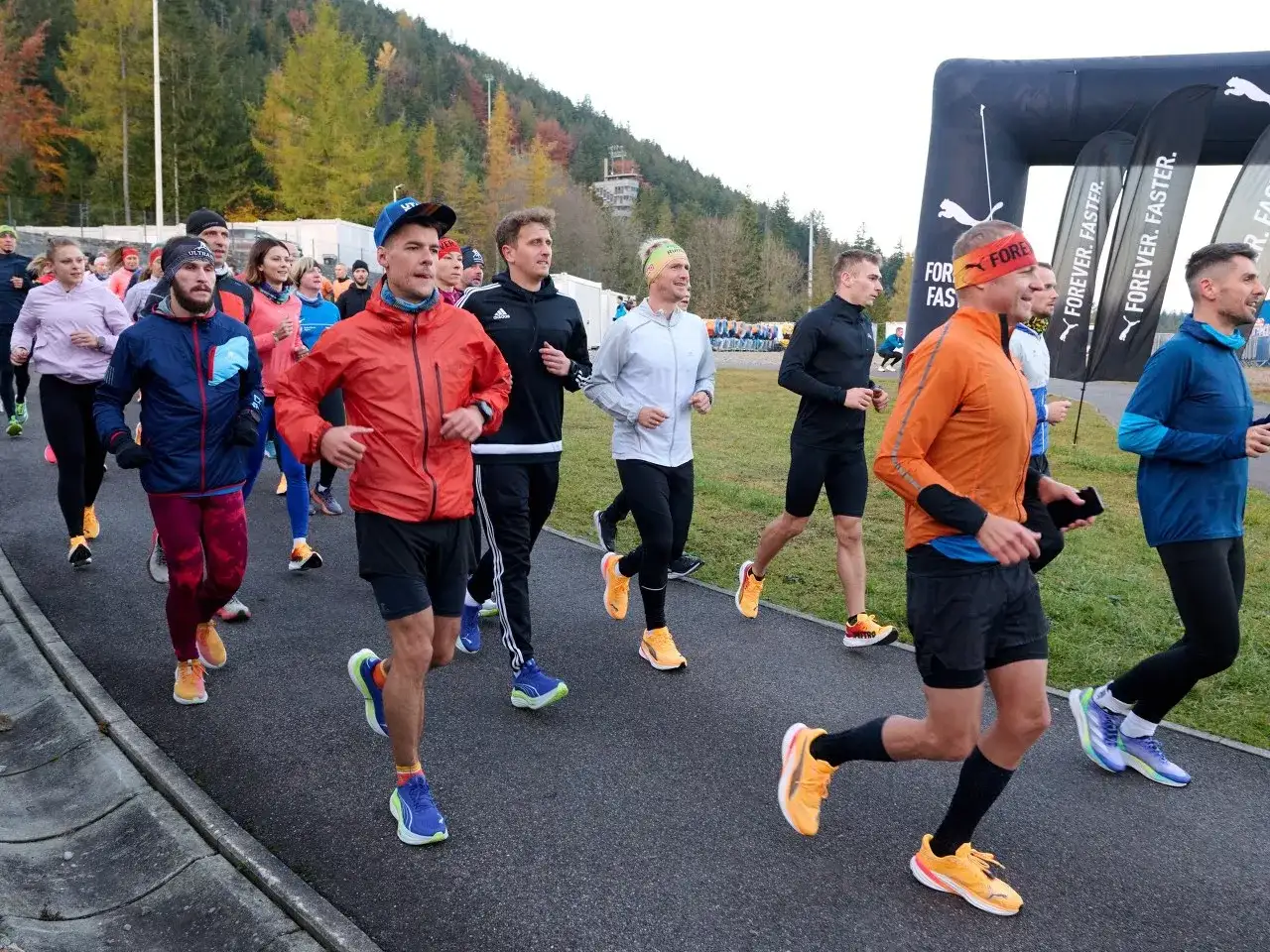 Grupa biegaczy w kolorowych strojach, jak z filmu o bieganiu, startuje na trasę. W tle las i bramka mety Puma.