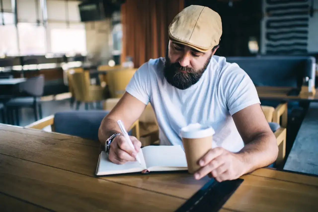 Mężczyzna w kaszkiecie pisze w notatniku, zastanawiając się, czy cytat to plagiat. Trzyma kubek kawy.
