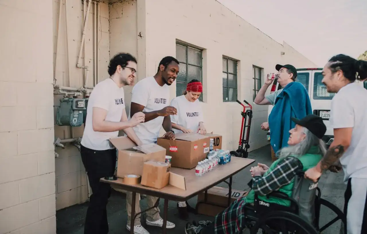 Grupa wolontariuszy w białych koszulkach pakuje paczki z pomocą, obok siedzi osoba na wózku inwalidzkim.