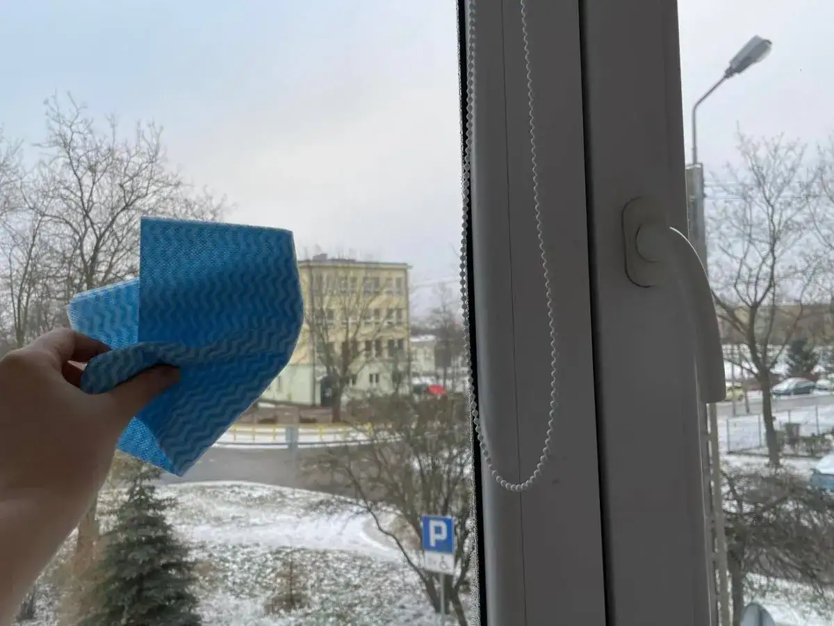 Ręka trzyma niebieską ściereczkę, gotową do mycia okna. Za szybą zimowy krajobraz z drzewami i budynkiem. Czy można myć okna na mrozie?