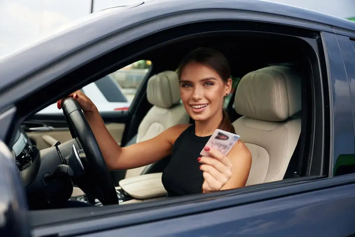 Uśmiechnięta młoda kobieta w samochodzie trzyma prawo jazdy. Zastanawiasz się, ile trwa kurs na prawo jazdy?