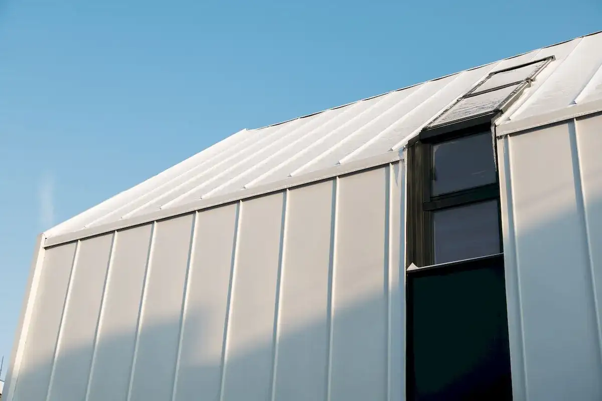 Nowoczesny budynek z białej blachy na rabek, pokryty śniegiem, z ciemnym oknem i świetlikiem.