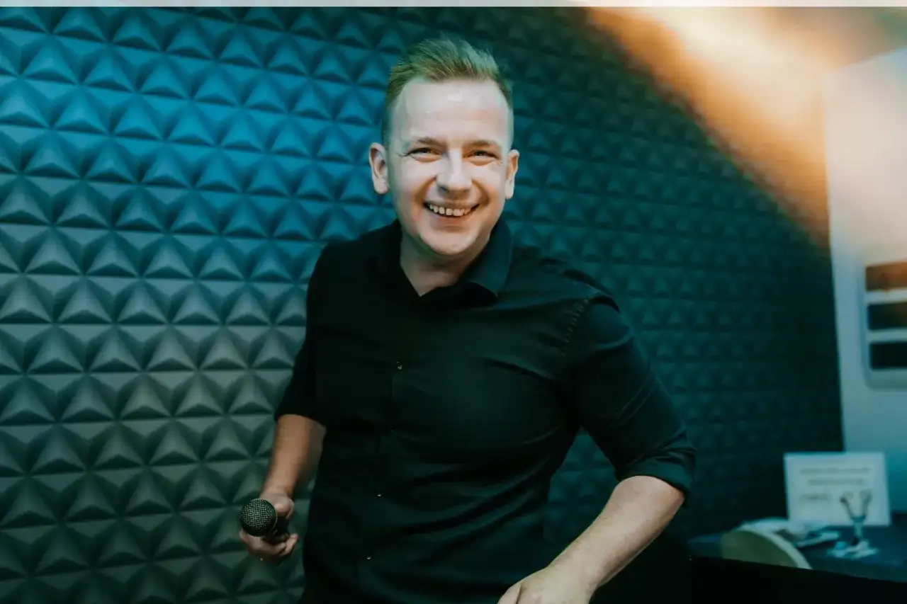 Uśmiechnięty DJ w czarnej koszuli trzyma mikrofon na tle niebieskiej, geometrycznej ściany.