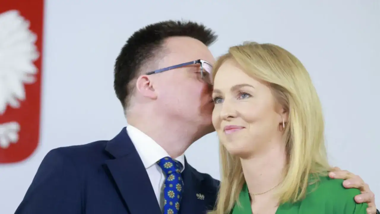 Szymon Hołownia szepcze coś do ucha Urszuli Brzezińskiej Hołowni. W tle godło Polski.