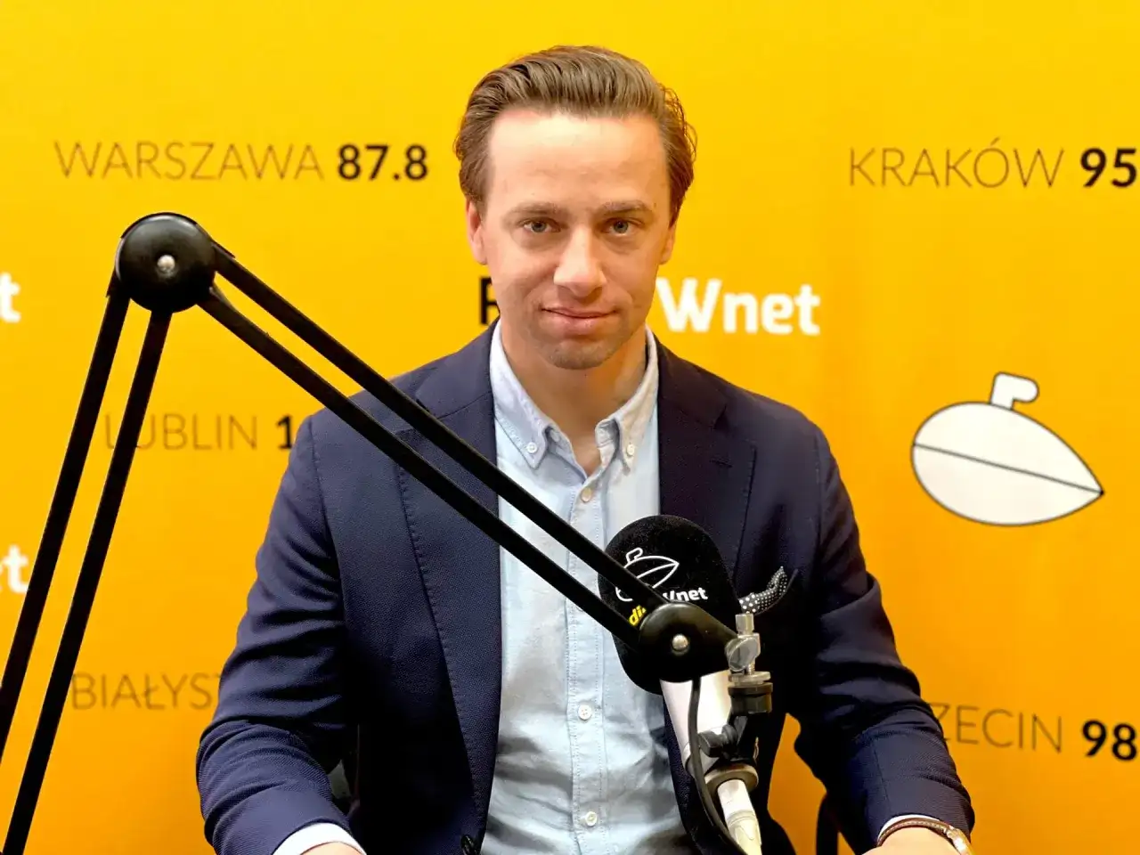 Krzysztof Bosak w garniturze przed mikrofonem na tle żółtej ściany z napisami.