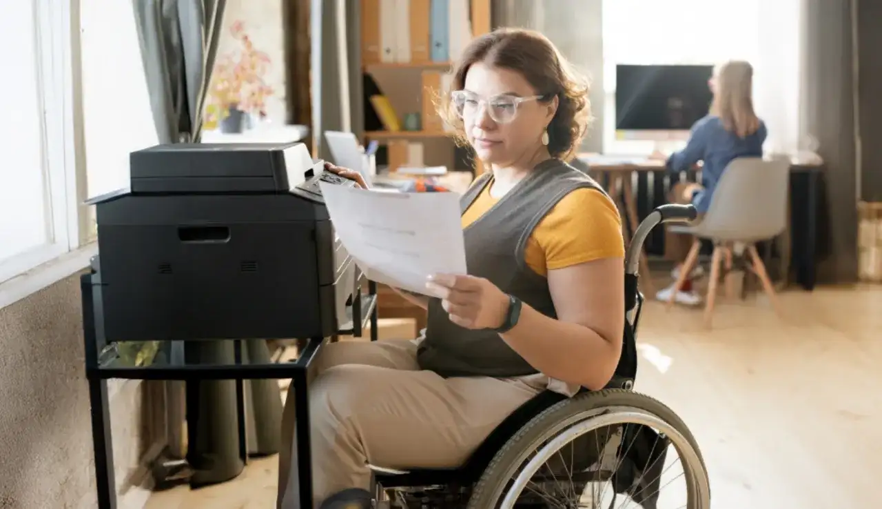 Kobieta na wózku inwalidzkim drukuje dokumenty. Zastanawia się, kto płaci asystentowi osoby niepełnosprawnej, gdy ona sama pracuje.