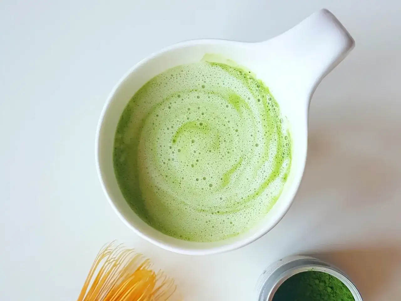 Zielony, spieniony napój matcha w białej miseczce, idealny jako alternatywa co zamiast kawy. Obok bambusowa miotełka i proszek matcha.