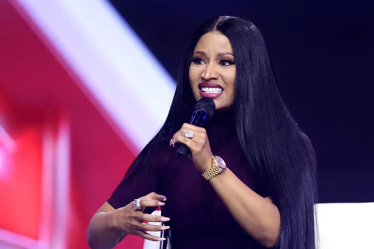 Nicki Minaj z mikrofonem, w ciemnej sukience, z długimi czarnymi włosami i biżuterią.