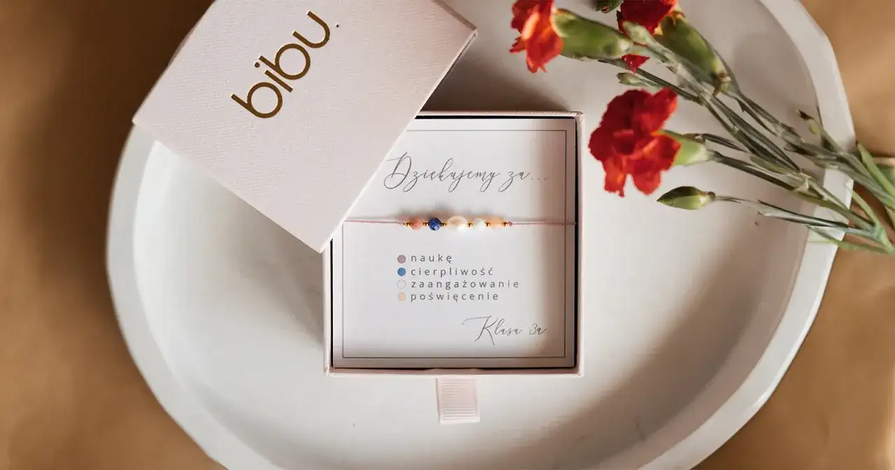 Otwarty różowy kartonik z bransoletką i napisem "Dziękujemy za...", obok czerwone goździki.