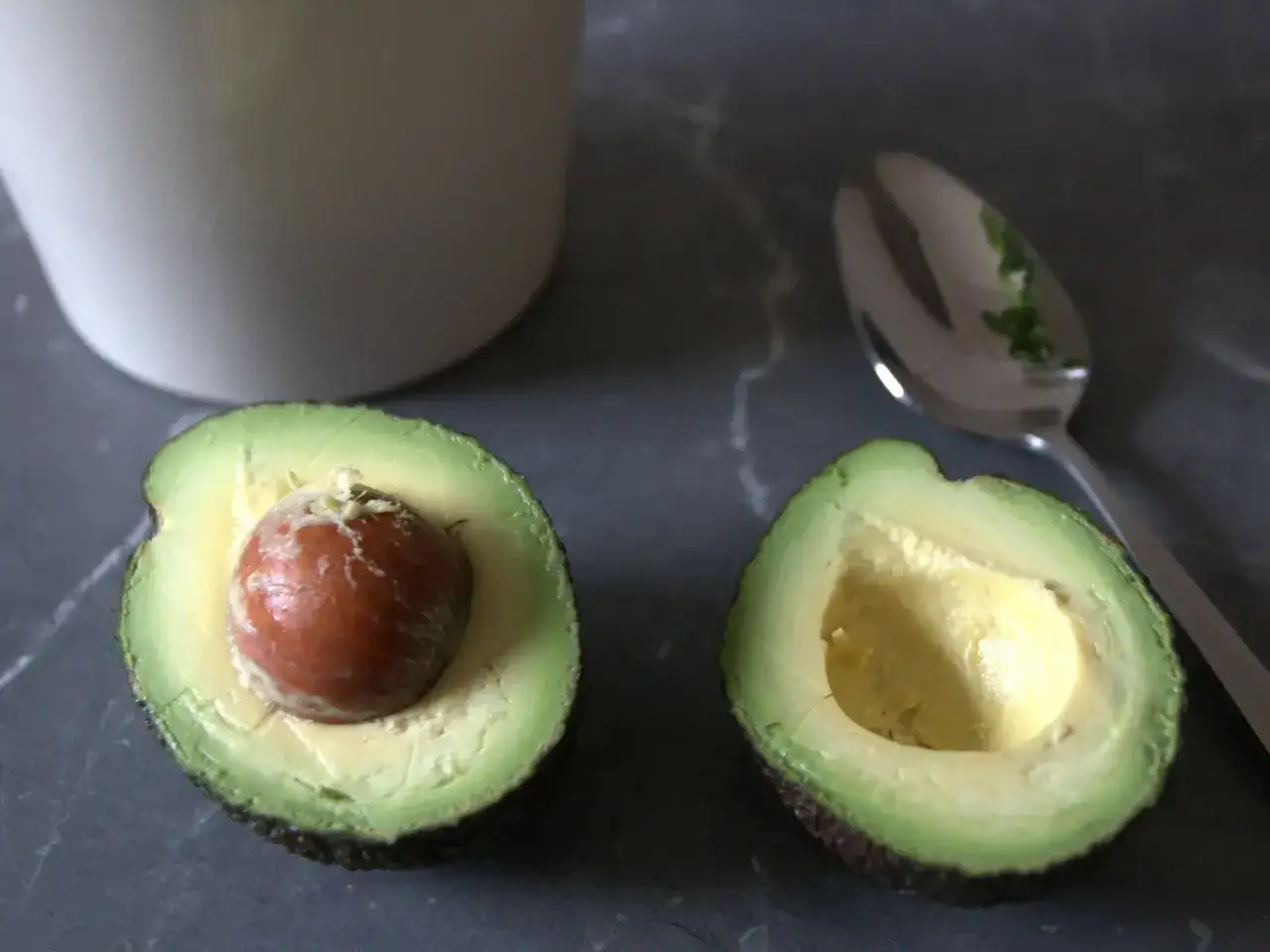 Dwa połówki awokado, jedna z pestką, druga pusta, obok łyżka. Prosty sposób, jak obierać awokado.