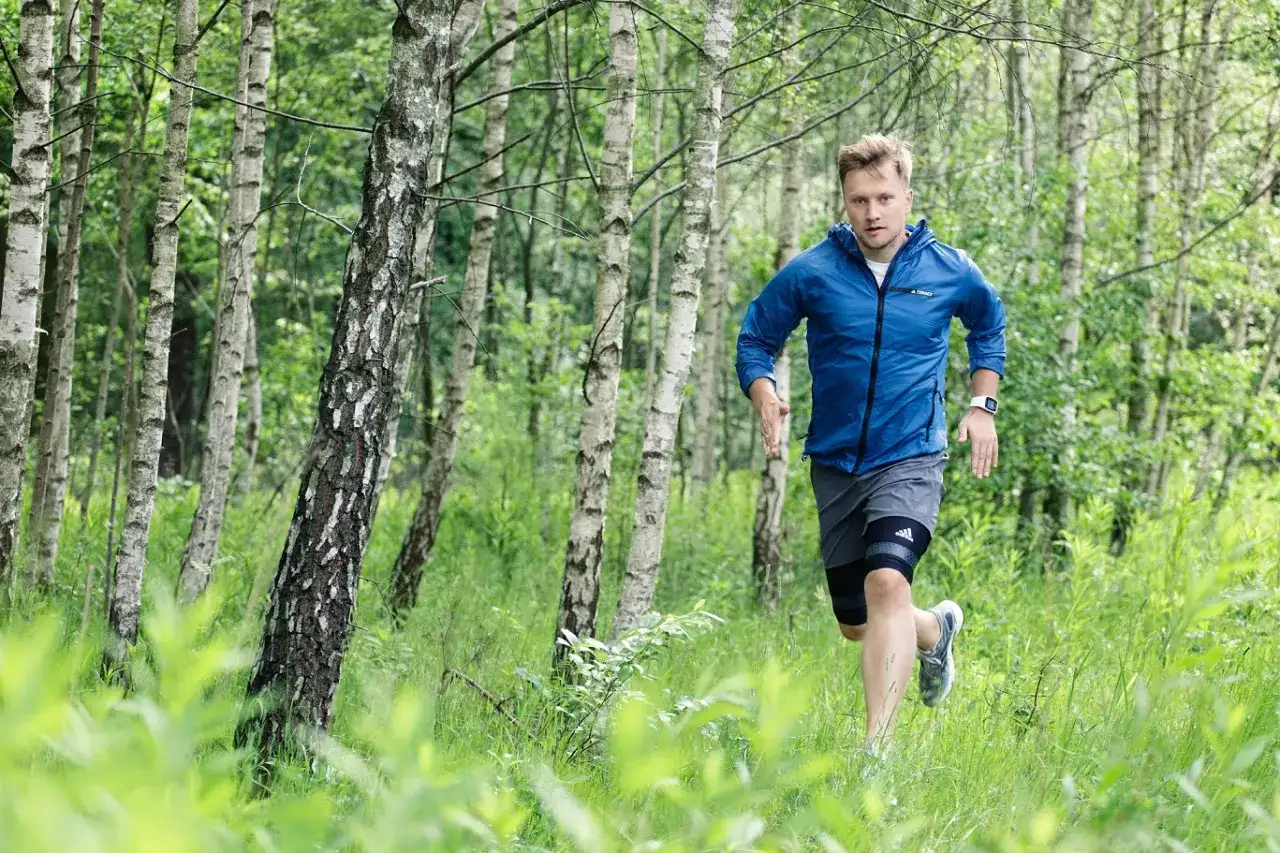 Mężczyzna w niebieskiej kurtce i szarej bluzie z szortami biegnie po lesie, otoczony zielenią i drzewami.