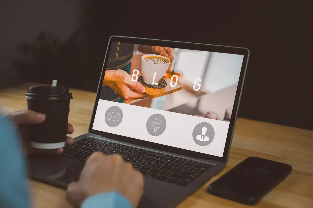 Osoba z kubkiem kawy pracuje przy laptopie, na ekranie którego widnieje napis "BLOG" i ikony.
