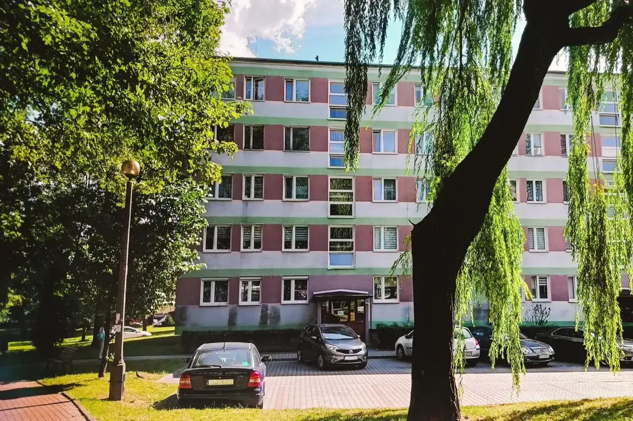 Blok mieszkalny z zielonymi i różowymi pasami, drzewa i zaparkowane samochody.