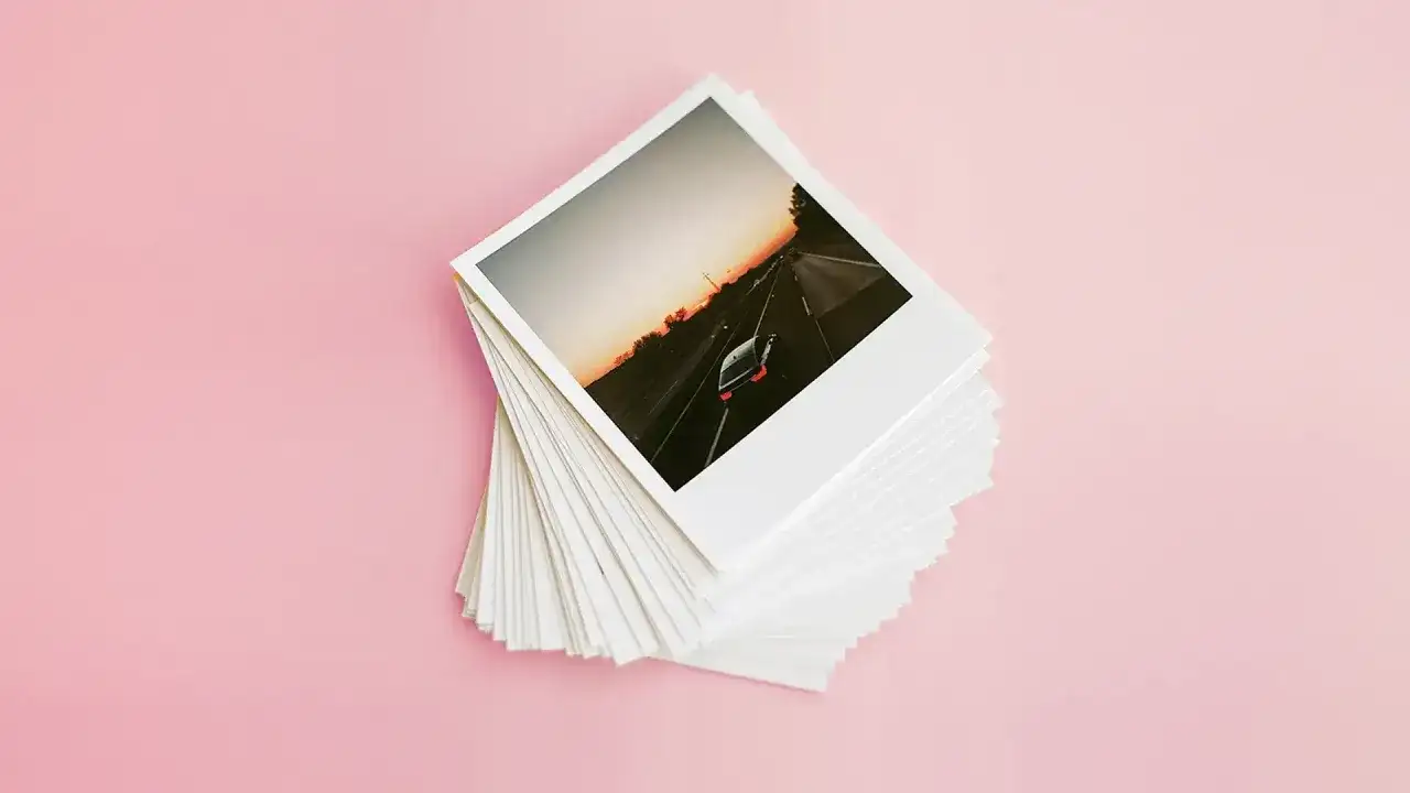 Stos odbitek z wakacyjnej podróży, idealny do wywoływania zdjęć w domu. Na wierzchu zdjęcie samochodu jadącego autostradą o zachodzie słońca.